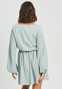 Svetlo modra obleka z dolgimi, zračnimi rokavi in cinčanim pasom. Ima teksturirano blago s črnimi pikami po celotni površini.