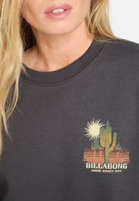 Donkergrijze sweatshirt met ribgebreide kraag, afgewerkt met een kleurrafische afbeelding van een zon en cactus, en de tekst "BILLABONG UNDER SUNSET SKIES."