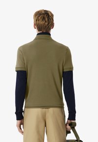 Man med kort blont hår som bär en olivgrön kortärmad skjorta över en marinblå långärmad topp, beige byxor och håller i ett grönt väskrem.