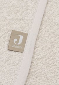 Witte katoenen handdoek met een textuur van badstof, voorzien van een beige label met witte tekst en een gestikte randaccent. Zachte en absorberende textuur.