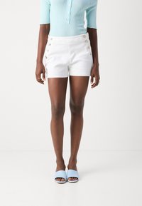 Pantalones cortos de algodón blanco con botones dorados en el lateral, combinados con una camiseta de canalé azul claro y sandalias azules sin talón.