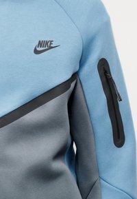 Μπλε και γκρι φούτερ Nike. Διαθέτει διαγώνιο μαύρο πάνελ, τσέπη με φερμουάρ στο μανίκι και μαύρο λογότυπο στο στήθος.