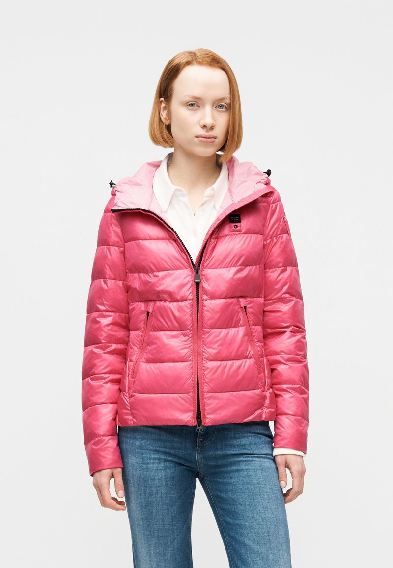 Νεαρή γυναίκα με κοντά κόκκινα μαλλιά που φοράει ροζ μπουφάν με φερμουάρ πάνω από λευκό μπλουζάκι και μπλε τζιν, στέκεται μπροστά σε απλό φόντο.