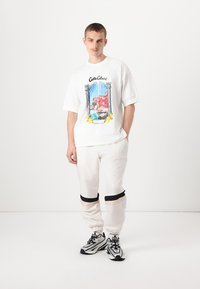 Camiseta blanca de gran tamaño con un diseño gráfico colorido que representa una escena costera. Combinada con pantalones jogger blancos que tienen acentos en negro y beige.