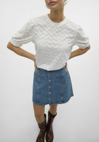 Hvid, kortærmet top med zigzag-mønster og hulmønster-detaljer, parret med en blå denimnederdel med frontknapper og lommer.