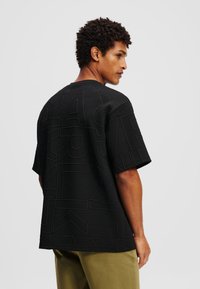 Tricou negru cu mânecă scurtă, cu litere și modele reliefate, țesătură texturată, guler rotund și croială lejeră, purtat cu pantaloni verde măsliniu.