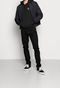 Černá bomber bunda přes černou mikinu, zkombinovaná s černými slim-fit džíny a černými teniskami s bílými podrážkami. Hladká tkanina, minimalistický design.