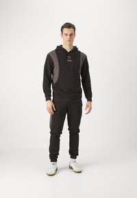 Felpa nera con cappuccio e pantaloni abbinati con accenti grigi; presenta una tasca frontale e un logo. Il materiale sembra morbido e testurizzato. Inclusa anche un paio di scarpe bianche.