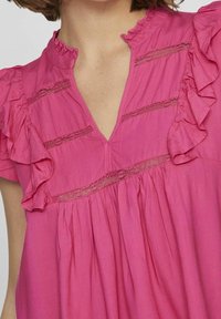 Rosa bomullsklänning med V-ringning, volangkrage och korta ärmar. Spetsdetaljer och veck förhöjer designen. Slät textur.