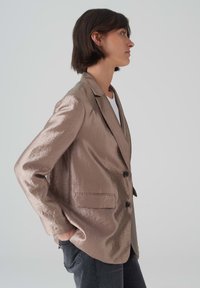Metallic beigefarbener Blazer mit glatter Textur, Ein-Knopf-Verschluss, Reverskragen und seitlichen Taschen, getragen über einem weißen Oberteil.