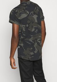Camiseta de manga corta hecha de algodón, con un patrón de camuflaje en verde oscuro y negro, con mangas enrolladas y un detalle de dobladillo curvado.