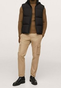 Veste matelassée noire avec capuche portée sur un pull côtelé marron, associée à un pantalon cargo beige avec poches latérales et des bottines noires.