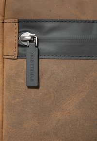 Texture in pelle marrone con una sezione con zip nera e un'etichetta nera con il marchio "STRELLSON". I dettagli di cucitura visibili migliorano il design.