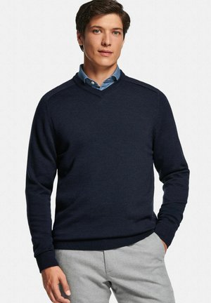 Pull col en V bleu marine en tissu doux, avec poignets et ourlet côtelés. Porté par-dessus une chemise bleu clair à col, de texture lisse.