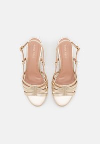 Korkeakantaiset, kultaiset slingback-kengät kiemuralla ja kiiltävällä pinnalla, joissa on kevyehkö pehmustettu pohja ja pyöreä varvas. Säädettävä nilkkahihna soljella.