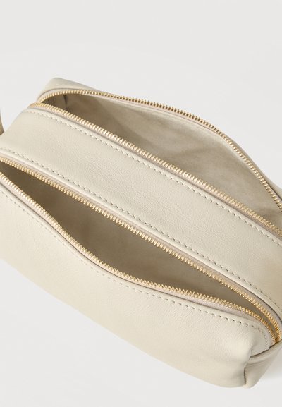 Μπεζ δερμάτινο τσαντάκι με δύο ανοιχτά χρυσά φερμουάρ που αποκαλύπτουν κενές εσωτερικές θήκες πάνω σε λευκό φόντο.