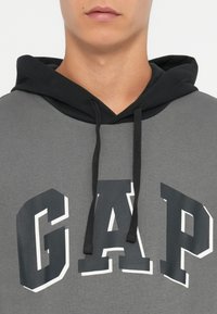 Hanorac gri cu glugă neagră, având un mare logo „GAP” în alb și negru. Materialul pare moale; sforile negre sunt vizibile.
