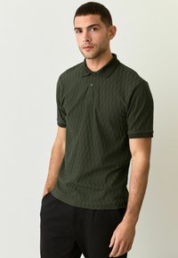 Camiseta polo de color verde oscuro con patrón texturizado, de mangas cortas y con un cuello clásico. Presenta un tapeta de dos botones y dobladillo con costura lateral.