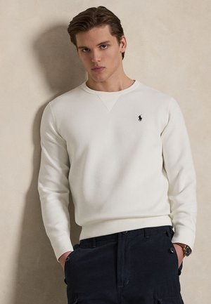 Mladi moški oblečen v belo dolg rokav pulover s majhnim črnim logotipom na prsih in temne hlače, stoji z rokami v žepih.