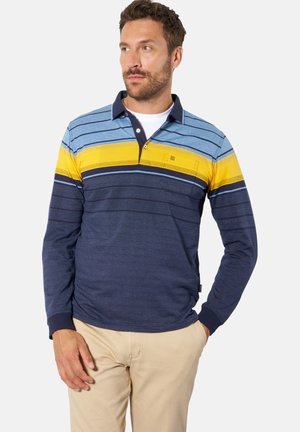 Mann mit Bart, der ein langärmeliges Poloshirt mit blauen, gelben und dunkelblauen Streifen und beigen Hosen trägt, steht mit einer Hand in der Tasche und blickt nach links.