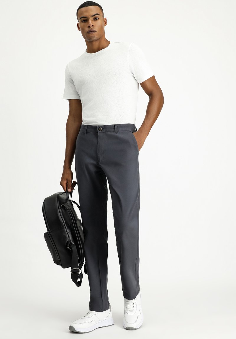 Herenoutfit met een aansluitend wit t-shirt met korte mouwen en donkergrijze broek. Het model houdt een zwarte rugzak vast en draagt witte sneakers.