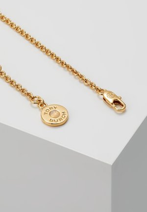 Collier chaîne ton or avec un pendentif circulaire gravé "TORY BURCH" et un fermoir mousqueton, présenté sur le bord d'une surface blanche.