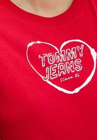 T-shirt rossa in cotone con grafica di un cuore bianco e la scritta "TOMMY JEANS" in caratteri grandi, con "Since 85" sotto. Maniche corte.