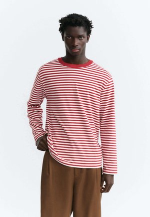 Giovane uomo con una camicia a righe rosse e bianche a maniche lunghe e pantaloni marroni, in piedi davanti a uno sfondo chiaro e uniforme.