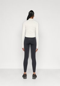 Λευκό μακρυμάνικο μπλουζάκι με εφαρμοστή γραμμή, σε συνδυασμό με μαύρα κολάν. Το μοντέλο φαίνεται από πίσω, φορώντας σκούρα αθλητικά παπούτσια.