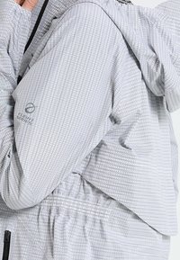 Osoba w jasnoszarej, teksturowanej kurtce Flight Series z elastycznym pasem i czarnym zamkiem, poprawiająca kołnierz w okolicy szyi.