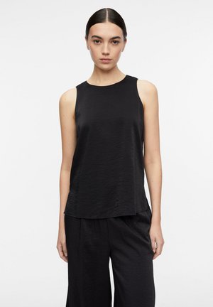 Donna con capelli scuri legati indietro, che indossa un top nero senza maniche e pantaloni neri larghi su uno sfondo bianco semplice.