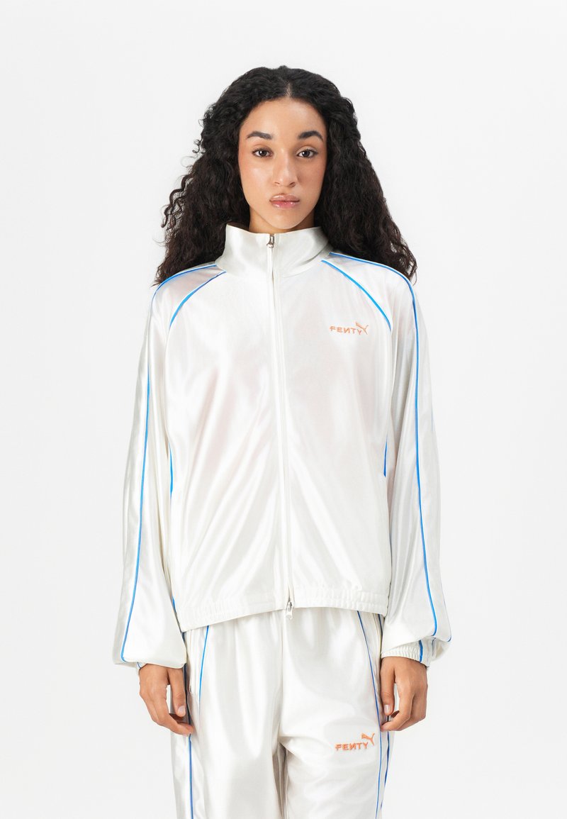 Top in tuta bianco con collare alto, caratterizzato da finiture blu e branding arancione "FENTY". Tessuto liscio e lucido con chiusura a zip.