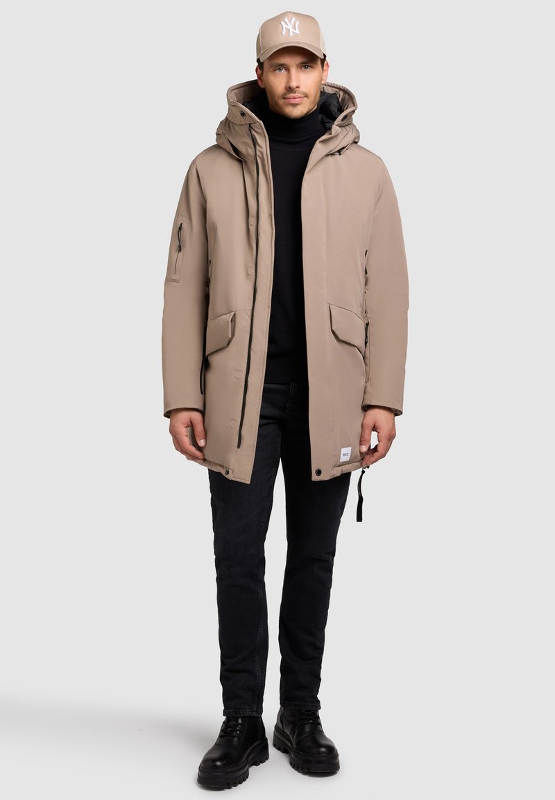 Parka beige con capucha, bolsillos frontales y acentos negros. Usado sobre un suéter negro de cuello alto y combinado con jeans oscuros y botas negras.