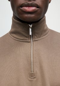 Μακροεντολή του κάτω μέρους του προσώπου και του λαιμού ενός ατόμου που φοράει ένα καφέ υψηλό γιακά με φερμουάρ και ασημένιο φερμουάρ.