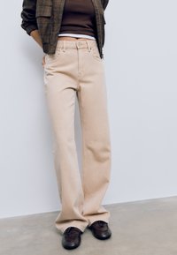 Bež široke traperice s blago teksturiranom tkaninom, s klasičnim gumbom i zatvaračem, prednjim džepovima i ravnim krojem.