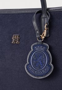 Navy színű filc bevásárlótáska, amely arany színű logóval és kék hímezett oroszlánmintás folttal rendelkezik, arany klipszel rögzítve.