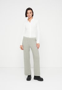 Jeune personne aux cheveux courts et foncés portant une chemise blanche boutonnée, un pantalon large gris à maille torsadée et des chaussures plateformes noires, debout devant un fond blanc.