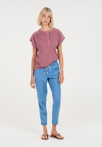 Lichte, mouwloze top in rozekleur met een knoopsluiting bij de hals, gecombineerd met blauwe, taps toelopende broek en bruin flip-flops.