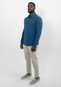 Blauwe lange mouwen polo met kraag, gecombineerd met lichtbeige slim-fit broek en grijze instappers, staand tegen een witte achtergrond.