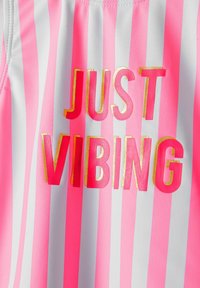 Maillot de bain avec des rayures verticales roses et blanches ; présente la phrase "JUST VIBING" en lettres roses et dorées ; matière lisse et extensible.