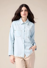 Light blue denim shirt with long sleeves, button-up front, two chest pockets, and frayed hem. Simplistic design with contrasting stitching.