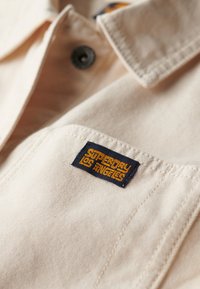Beige skjortekrave med en mørk marineblå lap, der har gul tekst "Superdry Los Angeles." Materialet ser struktureret ud med synlige søm.