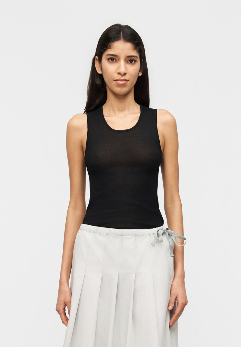 Νέα γυναίκα με μακριά σκούρα μαλλιά που φοράει μ αύρο αμάνικο τοπ και ανοιχτό γκρι πλισέ φούστα με φιόγκο στο πλάι.