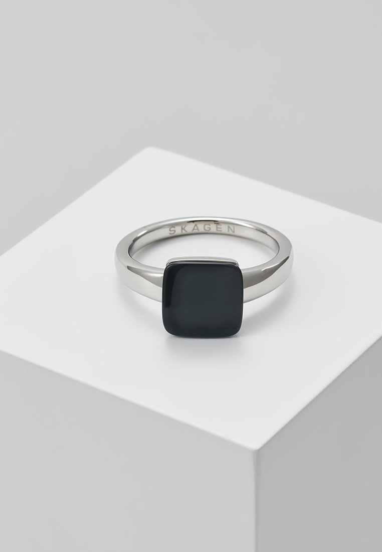 Bague en argent avec une pierre carrée noire polie montée sur le dessus, exposée sur un socle blanc avec "SKAGEN" gravé à l'intérieur de l'anneau.