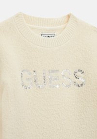 Pulover crem, cu un finisaj texturat, având un logo "GUESS" din paiete argintii. Guler ribat și țesătură moale.