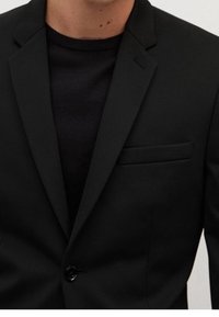 Mustapuku, joka on valmistettu sileästä kankaasta ja jossa on yksittäinen nappikiinnitys, lovetut kaulukset ja rintatasku. Yhdistetty mustan villapuseron kanssa.