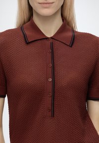 Femme portant un polo bordeaux texturé à manches courtes avec des bordures et des boutons noirs, montrée du torse au menton devant un fond uni.