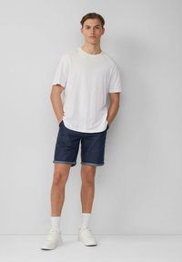 Weißes Baumwoll-T-Shirt, lockere Passform, kombiniert mit dunklen Jeansshorts mit umgeschlagenen Säumen. Weiße Sneaker und knöchellange Socken runden den Look ab.