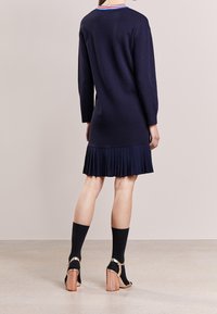 Abito maglione blu navy con orlo plissettato, caratterizzato da un collo rotondo con un bordo rosso, bianco e blu. Indossato con calze nere e sandali con tacco metallici.