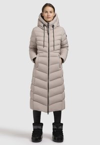 Long, light beige padded coat with a high collar and drawstring hood. Features zip closure, side pockets, and a chevron quilt pattern. Black footwear.